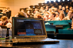 Ein Laptop steht auf einem Tisch, im Hintergrund ein Hörsaal mit vielen Menschen.