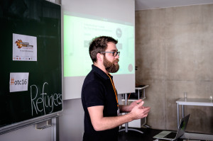 Ein junger Mann mit Bart erklärt vor einer Leinwand, auf der eine Präsentation projiziert wird, etwas.